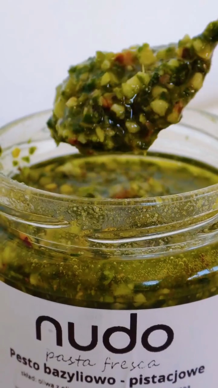 Pesto bazyliowo-pistacjowe NUDO na łyżce nad otwartym słoikiem, z widocznymi kawałkami składników.